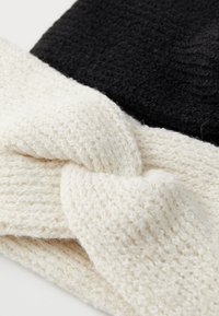 Cappello lavorato a maglia con sezione superiore nera e fascia intrecciata bianca; presenta un design attorcigliato; texture morbida con spessore del filato variabile.