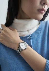 Relógio de metal prateado com um mostrador branco e detalhes azuis, usado no pulso. A pessoa que o usa está vestida com um suéter azul com uma gola alta clara por baixo.