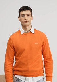 Oranje crewneck sweater met een textuur gebreid ontwerp, gedragen over een witte blouse met kraag. Heeft een subtiel logo op de borst.