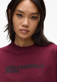 Burgundi värvi sinine dressipluus, millel on ümar kaelus ja musta värvi trükitud tekst "KARL LAGERFELD JEANS" ees. Pehme kangas ja lõdva disainiga.