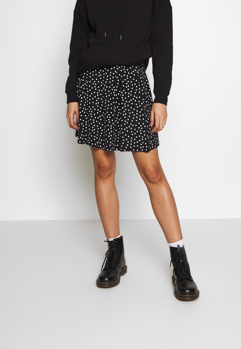 Persona vestida con sudadera negra, falda de lunares en blanco y negro, calcetines blancos y botas negras con cordones, de pie contra un fondo blanco.