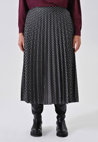 Schwarz-weißer plissierter Rock mit geometrischem Muster, aus leichtem Stoff mit glatter Textur. Getragen mit hohen schwarzen Stiefeln.