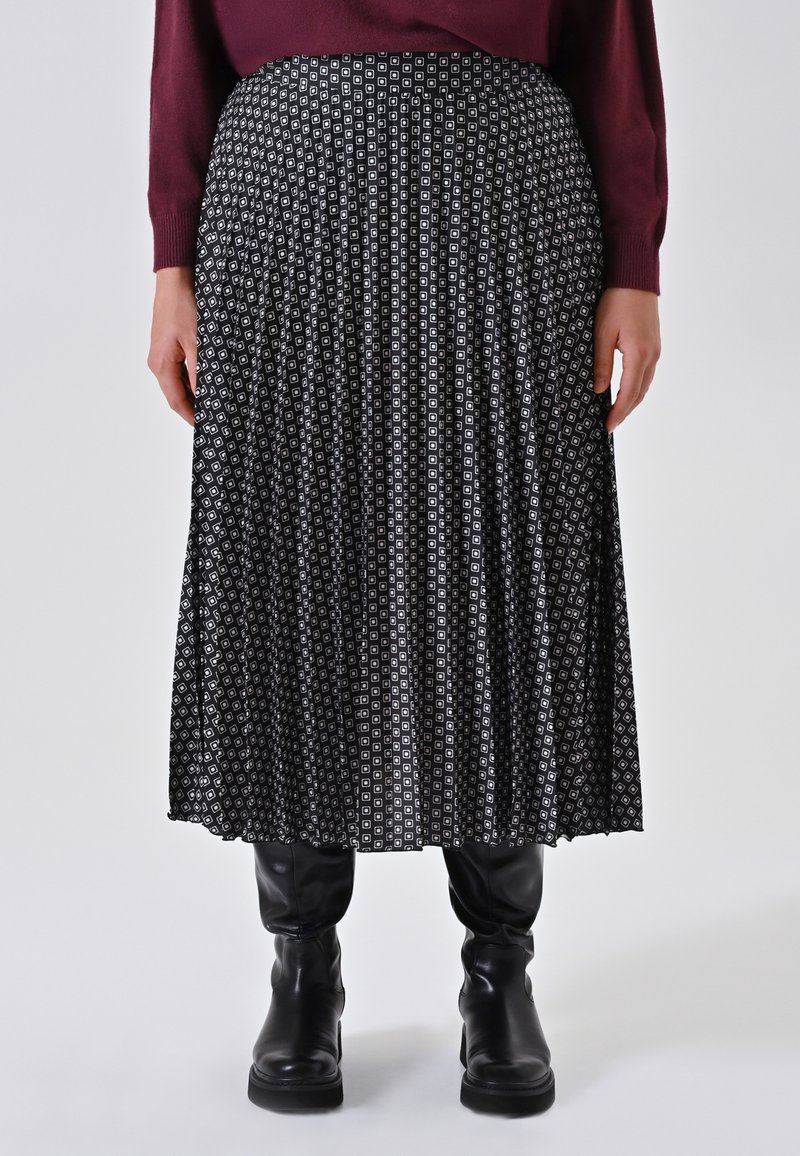 Schwarz-weißer plissierter Rock mit geometrischem Muster, aus leichtem Stoff mit glatter Textur. Getragen mit hohen schwarzen Stiefeln.