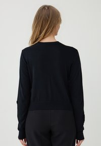 Frau mit langen blonden Haaren, die einen schwarzen langärmeligen Pullover und schwarze Hosen trägt, von hinten vor einem schlichten Hintergrund gezeigt.