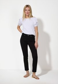 Weißes Oberteil mit puffärmel und runder Ausschnitt, kombiniert mit schwarzen Slim-Fit-Jeans und goldenen Sandalen. Der Hintergrund ist schlicht weiß.