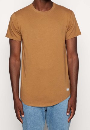 Homme portant un t-shirt marron uni à manches courtes associé à un jean bleu clair, sur un fond uni.