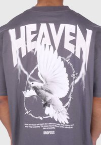 Tricou gri cu un text alb mare "HEAVEN" și o grafică cu o porumbel în cadrul unui cerc de sârmă ghimpată, având text suplimentar dedesubt, cu font mai mic.