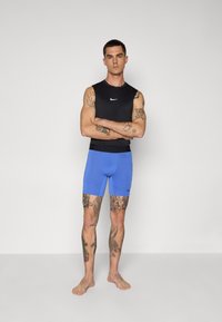Svart ermeløs topp med en hvit logo, kombinert med blå treningsshorts. Viser tatoveringer på underarmene og bena, barføtt foran en ensfarget bakgrunn.