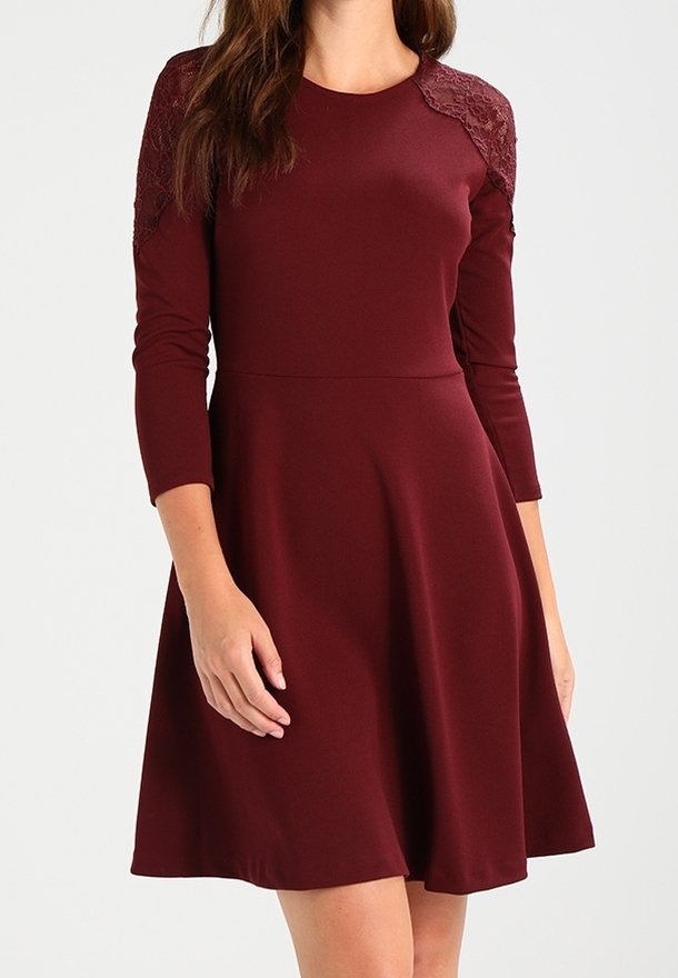 Femme portant une robe bordeaux mi-longue à manches longues avec des détails en dentelle sur les épaules et une taille cintrée.