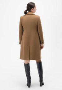 Bruin wollen cappot met een op maat gemaakt ontwerp, voorzien van een rugventilatie en lange mouwen, gecombineerd met zwarte leren kniehoge laarzen.