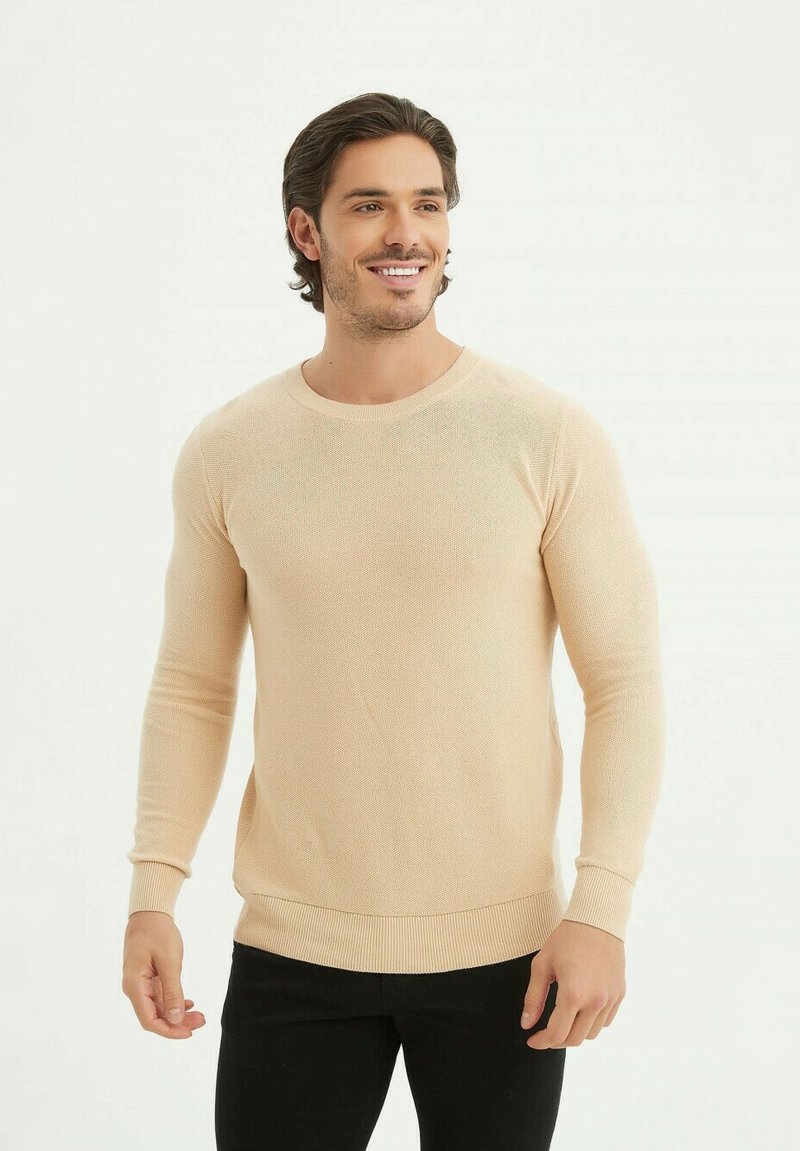 Pull tricoté beige clair avec un col rond, des manches longues, et des bords côtelés, associé à un jean noir ajusté.