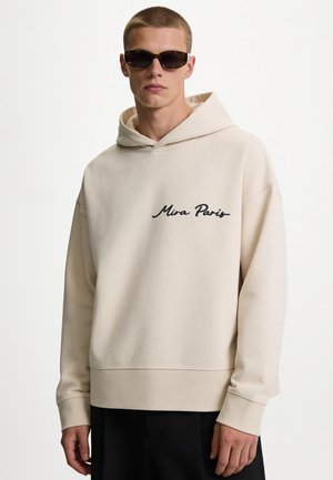 Junger Mann mit Schildpatt-Sonnenbrille und beigem Hoodie mit dem Text "Mira Paris" vorne, kombiniert mit schwarzen Hosen.