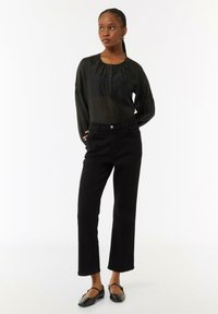 Schwarze Bluse mit gerafften Schultern und Knöpfen am Ausschnitt; kombiniert mit hochgeschnittenen, schwarzen, geraden Jeans und flachen, schwarzen Schuhen.