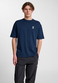 Marinblå kortärmad t-shirt i mjukt tyg, med en vit chevronlogotyp på bröstet, kombinerad med svarta byxor.