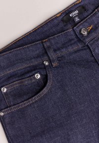 Tmavě modré džíny z denimu s oranžovým prošitím, obsahující přední kapsu, kovové zapínání na knoflík a štítek s nápisem "VERSUS."