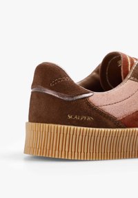 Bruine suède sneaker met een gestructureerde bovenkant, contrasterende oranje stof en een ribbelige gumzool. Beschikt over een gouden logo op de hiel.