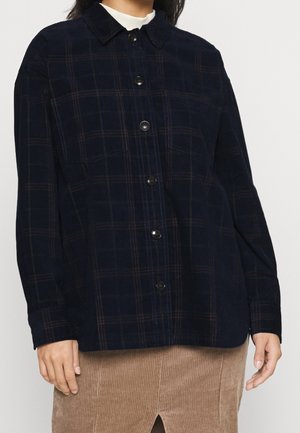 Persoon draagt een donker geruit overhemd met borstzakken en een lichtbruine corduroy rok met een voorste split.