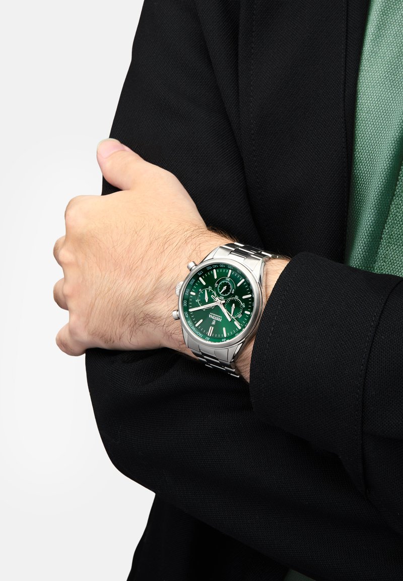 Montre en acier inoxydable argenté avec un cadran vert, dotée de deux sous-cadrans et d'accents argentés. Portée sur un poignet avec une manche de veste noire.