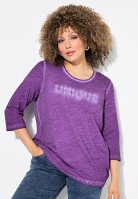 Lila, topp med trekvartsärmar och rund halsringning, med en textdesign i struktur som säger "UNIQUE" och tillverkad av mjukt, lätt material.
