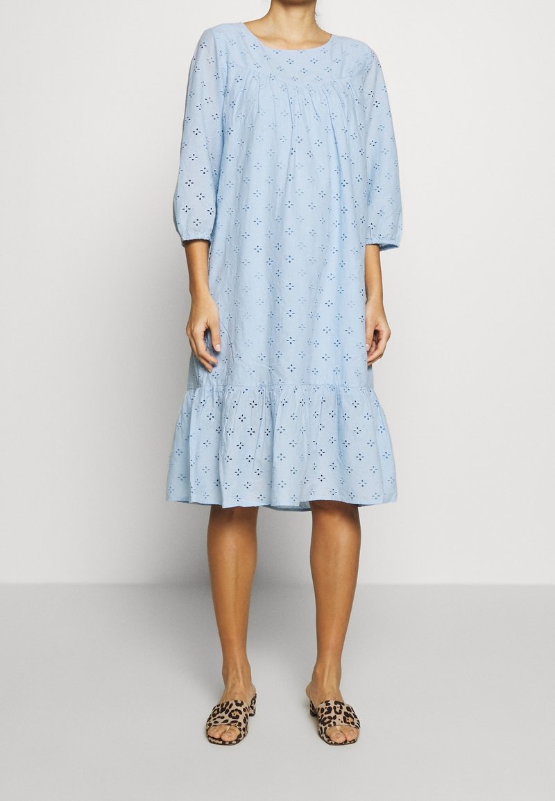 Femme portant une robe en eyelet bleu clair avec des manches 3/4 et un ourlet volanté, assortie à des sandales à enfiler ouvertes à l'avant à imprimé léopard.