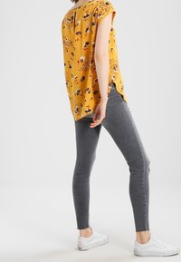 Persona che indossa una blusa corta a maniche corte gialla con fiori, jeans skinny grigi con orli sfrangiati e sneakers bianche, in posa di lato su uno sfondo bianco.