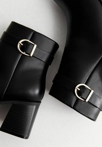 Bottines noires en cuir avec un bout arrondi et un talon chunky. Présente une accent en boucle dorée et une texture lisse.