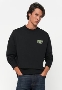 Νεαρός άνδρας με σκούρα μαλλιά, φόρεμα μαύρης μπλούζας με το λογότυπο "VERY CALVIN KLEIN" και μπλε τζιν, με τα χέρια στις τσέπες, κοιτάζοντας προς το πλάι.