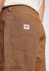 Pantalones de trabajo de algodón marrón que cuentan con un bolsillo trasero con una etiqueta blanca que dice "Lee Workwear". La textura es suave y tiene un ligero brillo.