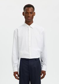 Λευκό πουκάμισο με μακριά μανίκια, κλασικό γιακά, κουμπιά μπροστά και μανσέτες, σε συνδυασμό με σκούρο μπλε παντελόνι. Λείο ύφασμα χωρίς σχέδια.