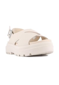 Lyse beige sandaler med en kraftig hvit såle, utskjæringsdesign og en sølvspenne på ankelremmen. Strukturert fotseng.