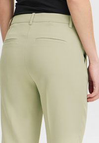 Svetlozeleni hlače iz gladkega materiala, z zadnjimi žepi in prilagojenim krojem. Pas ima zanke za pas za dodatne podrobnosti.