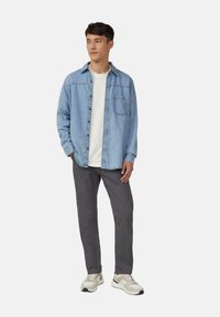 Jonge man in een lichtblauwe spijkerjack over een wit shirt, donkergrijze broek en witte sneakers, staand met één hand in zijn zak.