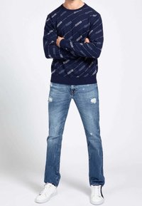 Jersey azul marino con el logotipo "GUESS" repetido en un patrón diagonal; combinado con jeans claros desgastados y zapatillas blancas. Estilo casual.