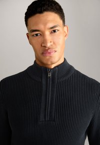 Marineblaue, gerippte Strickpullover mit hohem Kragen und halbem Reißverschluss. Der Stoff hat eine strukturierte Oberfläche und eine figurbetonte Silhouette.