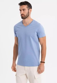 T-shirt di cotone azzurro chiaro con scollo a V, maniche corte e design aderente. Indossato con pantaloni cargo bianchi. Texture liscia, dettagli minimi.
