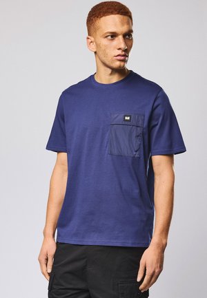 Jeune homme aux cheveux roux courts portant un t-shirt bleu avec une poche poitrine bleu foncé et un pantalon cargo noir, regardant sur le côté.