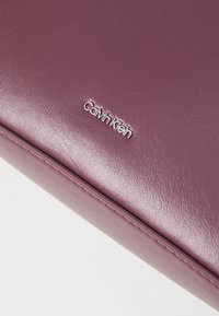 Calvin Klein kiegészítő, texturált lilás bőrből, fényes logó díszítéssel. Simára csiszolt felület, finom varrási részletekkel.