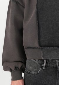 Sudadera oversized en mezcla de algodón en gris oscuro y negro. Presenta puños fruncidos y bordes deshilachados, combinada con jeans negros desgastados.
