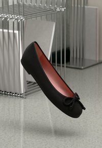 Sapatilha preta com textura suave, detalhe de laço na frente e interior vermelho contrastante. Design minimalista, bico arredondado.