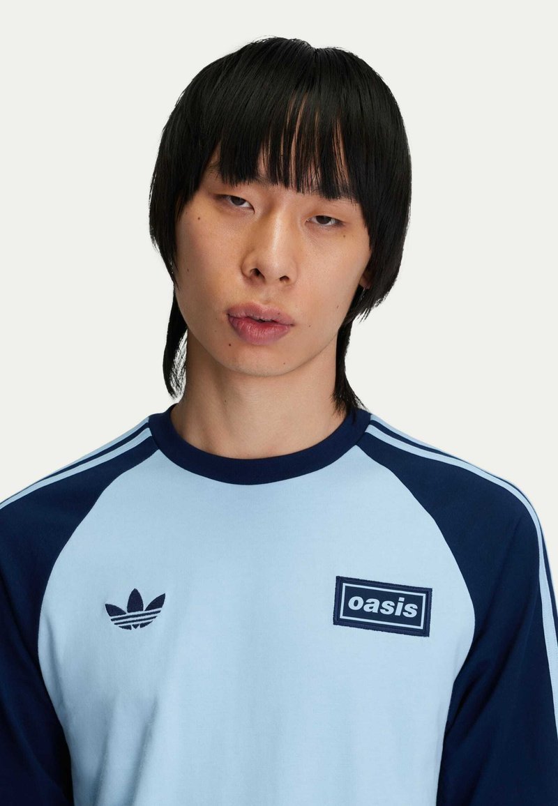 Camicia a maniche lunghe azzurro chiaro e blu navy con scollatura rotonda, dotata di un logo ricamato e strisce sulle maniche. Tessuto morbido al tatto.