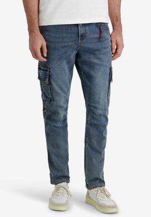 Man wearing slim-fit blue denim cargo jeans with side flap pockets and white sneakers, standing against a plain background.