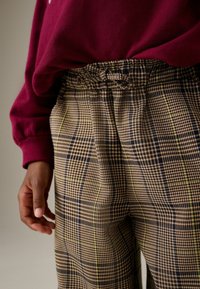 Pantaloni a scacchi marroni con accenti verdi e blu, dotati di una vita elastica e cordino. Il tessuto ha un aspetto testurizzato e intrecciato.