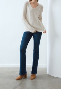 Krämfärgad kabelstickad tröja med V-ringning, kombinerad med mörkblå utsvängda jeans och ljusbruna klackade ankelskor på en slät betonggolv.