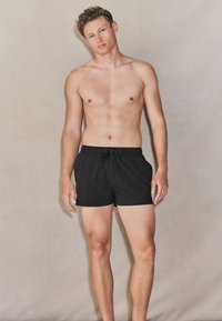 Mustad ujumispüksid kerge kangaga, millel on elastne vöökoht ja nöör. Mudel seisab paljajalu neutraalse tausta ees.