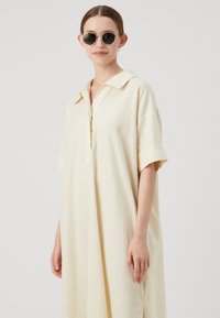 Abito corto di un leggero giallo chiaro realizzato in lino, con collo a camicia e maniche corte. Presenta una vestibilità ampia e cuciture laterali visibili.