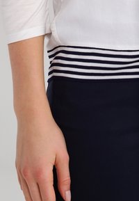 Weißes Oberteil mit drei Viertel langen Ärmeln und einem navy-blauen gestreiften Rippbund, kombiniert mit einer dunkelblauen Hose. Glatte Textur, taillierte Passform.