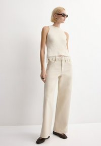 Mujer rubia con gafas de sol oscuras, top de punto sin mangas beige con borde de encaje, pantalones anchos beige, zapatos tejidos negros, de pie contra una pared blanca.