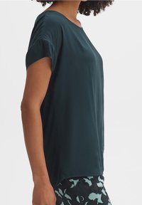 Krátké rukávy tmavě petrolejového topu s kulatým výstřihem, hladkou texturou a bočními rozparky, nošený přes vzorované černé a petrolejové legíny.