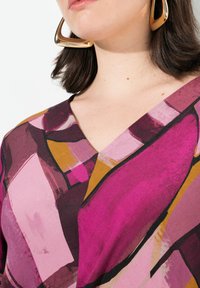 V-Ausschnitt-Oberteil mit einem lebhaften abstrakten Muster in Pink-, Lila- und Goldtönen; glatte Textur; kombiniert mit goldenen geometrischen Ohrringen.