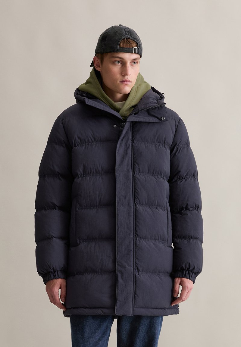 Gestepptes Dunkelblaues Jackett mit Kapuze, ausgestattet mit einem Frontreißverschluss, elastischen Bündchen und einer lockeren Passform, über einem olivgrünen Hoodie getragen.